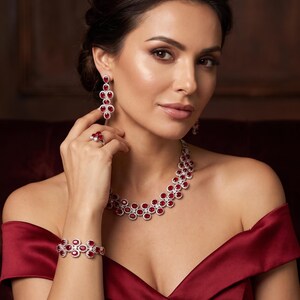 Puede incluir: Un conjunto de joyería compuesto por un collar, una pulsera, un anillo y pendientes colgantes. Cada pieza presenta piedras ovaladas de color rojo intenso rodeadas por pequeñas piedras claras en un marco de metal plateado, creando un patrón floral.