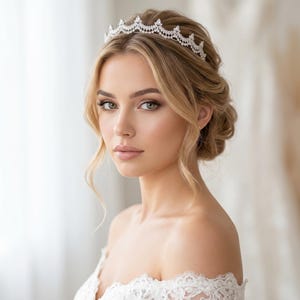 May include: A silver-toned tiara featuring a series of clear, faceted stones arranged in a repeating pointed, crown-like pattern. The headpiece is worn across the top of the head, resting against blonde hair styled in an updo with loose strands.