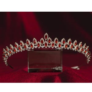 May include: A silver tiara adorned with red and clear gemstones. The tiara showcases red marquise-cut stones interspersed with clear, round-cut stones. It is presented on a clear, rectangular stand against a red backdrop.
