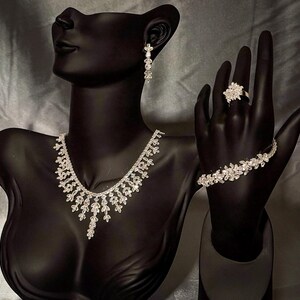 May include: A silver-toned jewelry set featuring a necklace with leaf-shaped crystal accents, a matching bracelet with floral-inspired crystal links, a cluster-style ring, and a single drop earring, all displayed on a black mannequin bust.