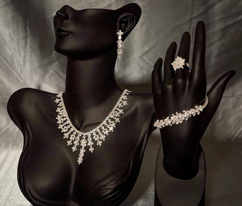 May include: A silver-toned jewelry set featuring a necklace with leaf-shaped crystal accents, a matching bracelet with floral-inspired crystal links, a cluster-style ring, and a single drop earring, all displayed on a black mannequin bust.