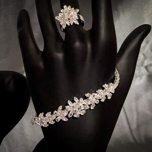 May include: A silver-toned bracelet and a matching ring, both featuring a cluster of clear, marquise-cut stones arranged in a floral pattern. The jewelry pieces are displayed on a black mannequin hand, highlighting the reflective, faceted surfaces.