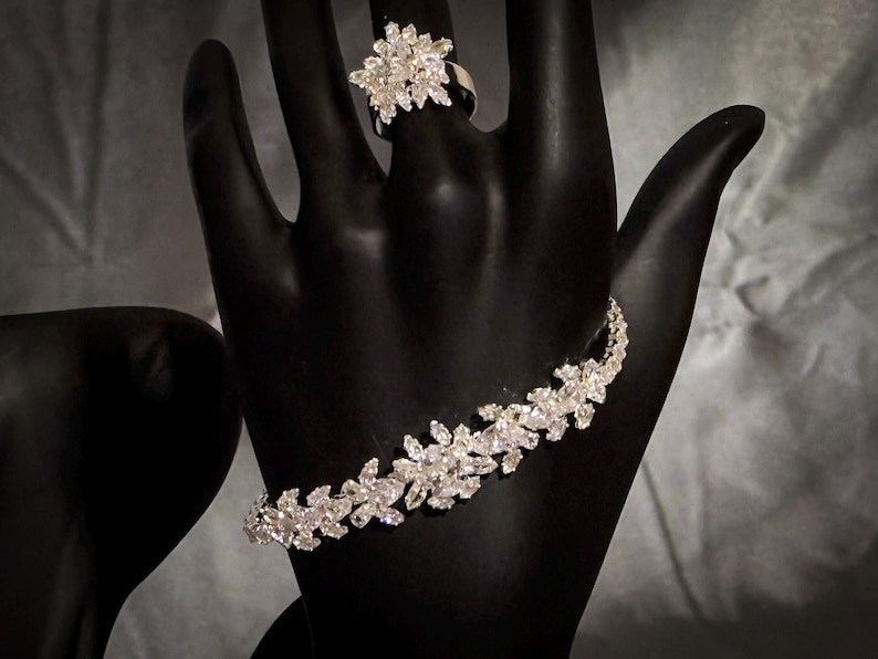 May include: A silver-toned bracelet and a matching ring, both featuring a cluster of clear, marquise-cut stones arranged in a floral pattern. The jewelry pieces are displayed on a black mannequin hand, highlighting the reflective, faceted surfaces.