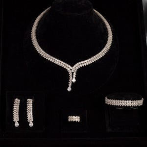 以下が含まれることがあります： ネックレス、イヤリング、ブレスレット、指輪がセットになったジュエリーセット。銀色の金属製で、多数の透明で輝く石で飾られています。ネックレスには、2つのぶら下がる石が付いたペンダントが付いています。