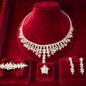 May include: A jewelry set consisting of a necklace, a bracelet, a ring, and a pair of drop earrings. Each piece features a silver-toned metal base adorned with clear, faceted stones arranged in a repeating leaf-like pattern throughout the set.