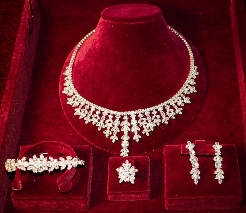 May include: A jewelry set consisting of a necklace, a bracelet, a ring, and a pair of drop earrings. Each piece features a silver-toned metal base adorned with clear, faceted stones arranged in a repeating leaf-like pattern throughout the set.