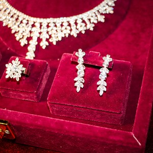May include: A jewelry set featuring a necklace, a ring, and a pair of drop earrings, all crafted with clear, faceted stones arranged in leaf-like patterns. The pieces are displayed on a deep red velvet surface with a gold-toned metal clasp.