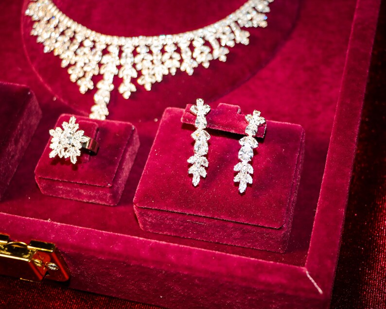 May include: A jewelry set featuring a necklace, a ring, and a pair of drop earrings, all crafted with clear, faceted stones arranged in leaf-like patterns. The pieces are displayed on a deep red velvet surface with a gold-toned metal clasp.