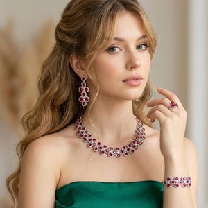 Może przedstawiać: Pasujący zestaw biżuterii składający się z naszyjnika, kolczyków, bransoletki i pierścionka. Zestaw zawiera czerwone kamienie szlachetne i małe, przezroczyste kamienie osadzone w srebrnym metalu. Kobieta na zdjęciu ma na sobie zielony top bez ramiączek.