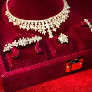 May include: A jewelry set featuring a silver-toned necklace, a bracelet, a ring, and a pair of drop earrings, all adorned with clear, faceted crystals in a floral-inspired pattern, displayed within a deep red velvet-lined presentation box.