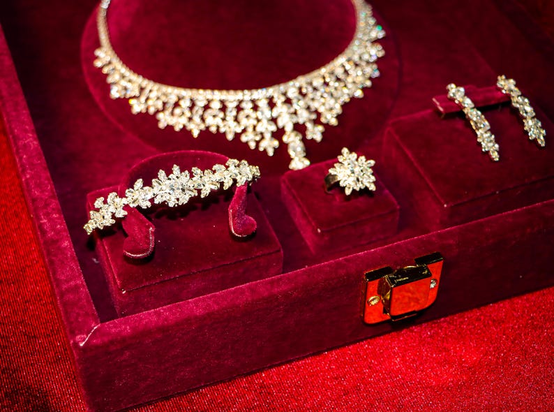 May include: A jewelry set featuring a silver-toned necklace, a bracelet, a ring, and a pair of drop earrings, all adorned with clear, faceted crystals in a floral-inspired pattern, displayed within a deep red velvet-lined presentation box.