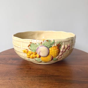 May include: A cream-coloured ceramic bowl with a textured basket-weave pattern on the exterior. The front features a raised, hand-painted relief design of assorted fruits, including yellow grapes, a purple plum, a yellow peach, and green leaves.