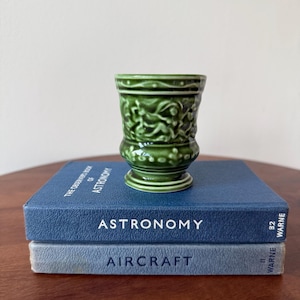 May include: A small, green ceramic cup with a detailed relief pattern. The cup is placed on top of two blue books titled "Astronomy" and "Aircraft". The cup has a pedestal base and a textured exterior.