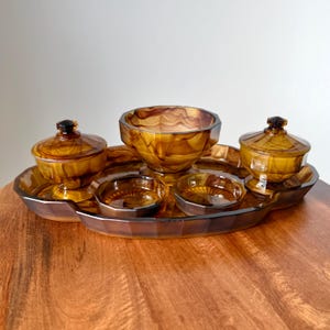 May include: An amber-coloured glass serving set. The set includes a large central bowl, two smaller lidded bowls, and several small dishes, all arranged on a matching tray. The glass has a marbled appearance, and the set is displayed on a wooden surface.