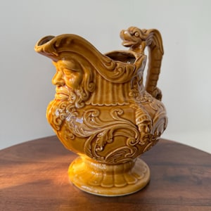 May include: A ceramic pitcher in shades of brown, featuring a detailed relief of a bearded face on the front. The handle is adorned with a dragon-like figure. The pitcher has a wide base and a spout, showcasing an antique aesthetic.