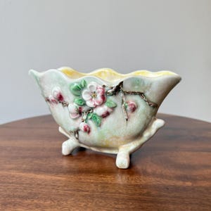 Könnte beinhalten: Ein Keramik-Pflanzgefäß mit gewelltem Rand und drei kleinen Füßen. Der Pflanztopf ist hellgrün mit erhabenen floralen und Astdetails in Rosa, Weiß und Grün. Der Innenraum ist gelb.