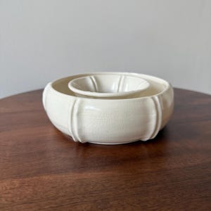 May include: Cream-coloured ceramic bowl set. The larger bowl has vertical ribbed accents and a crackled surface. A smaller, fluted bowl sits inside. The set is on a dark wooden surface.