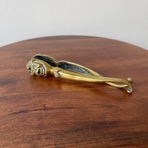 May include: A brass nutcracker featuring a sculpted human face on the head section with a textured, elongated handle. The metal tool has a polished gold-toned finish and a hinged mechanism designed for cracking nuts, showing visible aged patina.
