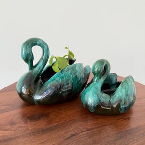 May include: Two ceramic swan planters with a glossy teal and black glaze. One planter holds a small green plant. The swans are displayed on a wooden surface. The planters are decorative and suitable for small plants.