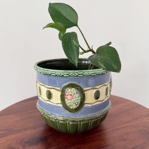 May include: A ceramic plant pot in blue, green, and cream. The pot has a floral design and contains a green leafy plant. The pot is approximately 15 cm tall and 15 cm in diameter. A decorative item for the home.