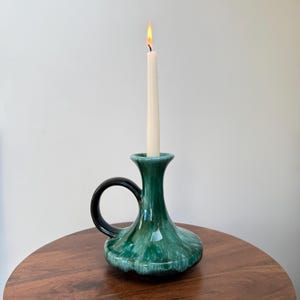 May include: A green ceramic candlestick with a black handle, holding a lit white candle. The candlestick has a flared base and a glossy finish. The candle is tall and slender, with a visible flame. The candlestick is on a wooden surface.