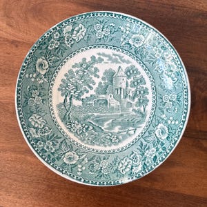 May include: A round, white and teal plate with a detailed floral border. The centre features a scenic illustration of a castle, trees, and a bridge. The plate is made of ceramic.