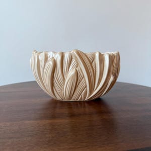 May include: A beige ceramic bowl with a textured, leaf-like design. The bowl has a rounded shape with a scalloped rim. The bowl is sitting on a dark brown wooden surface.