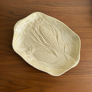 Könnte beinhalten: Eine hellgelbe, längliche Keramikservierplatte mit einem detaillierten geprägten Design von zwei Maiskolben. Die Platte hat eine wellenförmige, organische Form und steht auf einer dunklen Holzoberfläche.