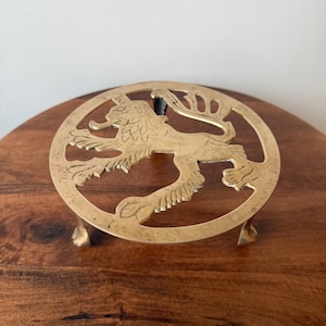 May include: A circular brass trivet featuring an openwork design of a rampant lion. The metal object has a polished gold-toned finish and is supported by three small, curved feet. The lion figure is centred within the circular frame.