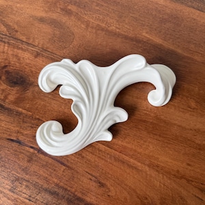 May include: A white ceramic wall hook featuring a glossy finish and an ornate, curved acanthus leaf design. The decorative hardware piece has a smooth, moulded surface with sweeping, fluid lines that extend outward into a prominent, curled hook shape.