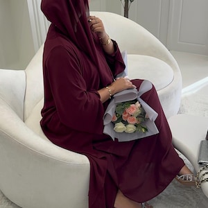 Puede incluir: Una persona con un vestido y un velo burdeos, sentada en una silla blanca, sosteniendo un ramo de flores. El vestido tiene mangas largas y un diseño fluido. El ramo incluye rosas blancas y rosas. La persona lleva pulseras doradas y sandalias.