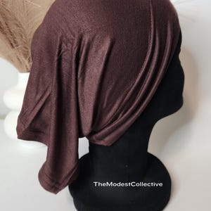 May include: A dark brown head covering, draped over a black mannequin head. The fabric has a subtle sheen and drapes softly. The text "TheModestCollective" is visible on the base.