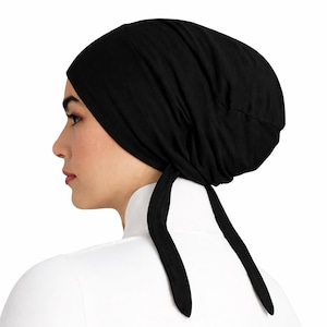May include: A black head covering with long ties, viewed from the side. The head covering is made of a soft fabric and is worn over the head. The ties hang down the back.