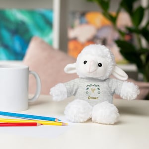 May include: A white plush lamb wearing a gray shirt with the name "Omar" and a green and gold design. The lamb has fluffy white fur and is sitting on a white surface. A white mug and colored pencils are in the background.