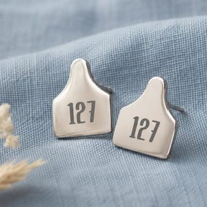 May include: A pair of silver-toned stud earrings shaped like livestock ear tags, each featuring the engraved number 127 in a dark, bold font. The earrings have a smooth, polished metallic finish and a standard post backing for secure attachment.