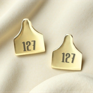 May include: Two gold-colored metal tags, each with the number "127" printed in dark gray. The tags have a curved top and a rectangular base. The tags are displayed on a cream-colored fabric.