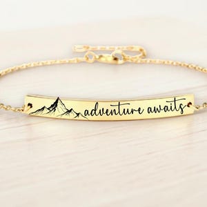 May include: A gold-colored bracelet with a rectangular bar pendant. The pendant features a mountain range graphic and the words "adventure awaits" in a script font. The bracelet has a delicate chain.