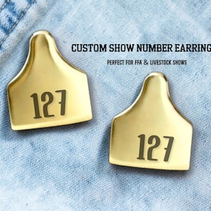 Könnte beinhalten: Zwei goldfarbene, individuelle Show-Nummer-Ohrringe mit der Nummer 127 in Dunkelbraun aufgedruckt. Die Ohrringe befinden sich auf einem hellblauen Jeans-Hintergrund. Der Text "CUSTOM SHOW NUMBER EARRINGS" steht über den Ohrringen.