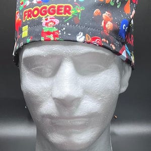May include: A black scrub cap featuring a colorful all-over print of various retro video game characters and logos, including the text FROGGER and BUBBLE BOBBLE. The fabric pattern displays multiple iconic figures in vibrant colors against a dark background.