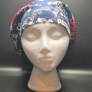 May include: A blue, red, and white patterned scrub cap featuring the text "PATRIOTS" and team logos. The cap is made of fabric and is designed to fit the head. The cap is displayed on a white mannequin head.