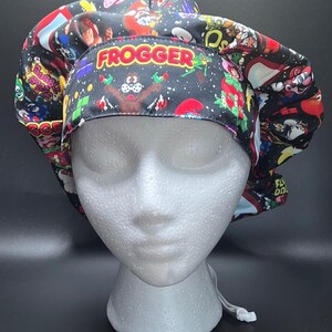 May include: A black scrub cap featuring a colorful, repeating pattern of classic 1980s arcade game characters and logos. The front band prominently displays the word FROGGER in yellow and red text, set against a dark background with pixelated graphics.