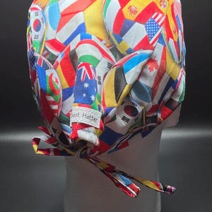 K&ouml;nnte beinhalten: Eine bunte OP-Haube mit einem Muster aus verschiedenen Nationalflaggen. Die Haube wird hinten mit einem passenden Stoffband gebunden. Das Etikett lautet "Best Hatter".