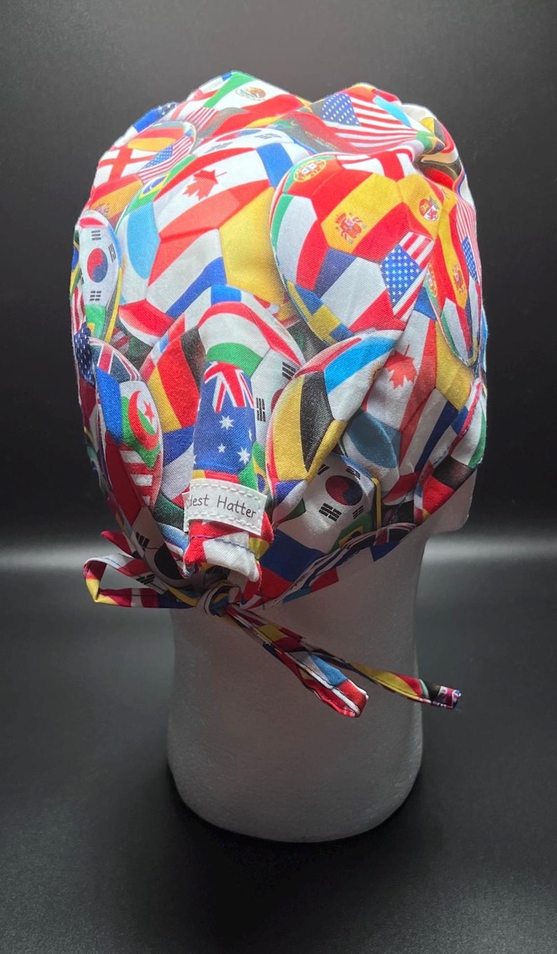 K&ouml;nnte beinhalten: Eine bunte OP-Haube mit einem Muster aus verschiedenen Nationalflaggen. Die Haube wird hinten mit einem passenden Stoffband gebunden. Das Etikett lautet "Best Hatter".