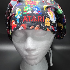May include: A black scrub cap featuring a colorful pattern of classic video game characters and the word "ATARI" in red. The cap is designed to fit a head and has a white drawstring. The overall theme is retro gaming.