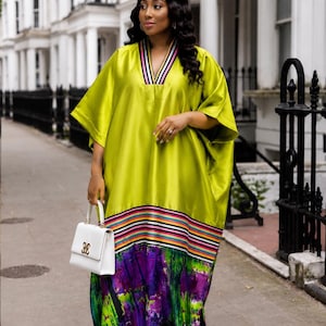 May include: A lime green kaftan dress featuring a V-neckline with black, white, and pink striped trim. The garment has wide sleeves and a horizontal striped band above a purple, green, and black abstract tie-dye pattern at the hemline.