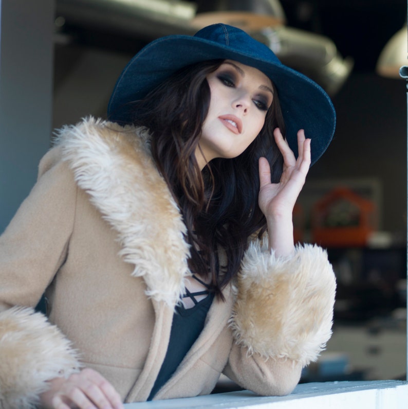 May include: A tan coat with a faux fur collar and cuffs. The coat is paired with a denim blue hat. The person in the image is wearing a black top.