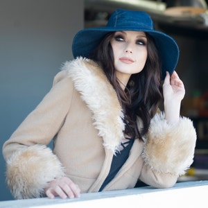May include: A woman wearing a denim blue hat and a tan coat with a faux fur collar and cuffs. The coat is buttoned and the woman is looking up and to the right.