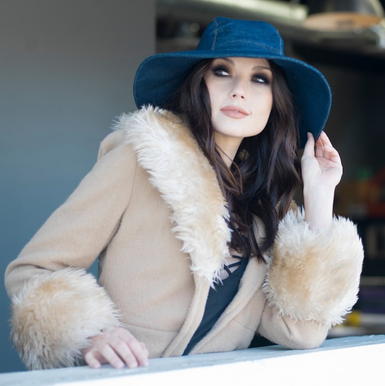 May include: A woman wearing a denim blue hat and a tan coat with a faux fur collar and cuffs. The coat is buttoned and the woman is looking up and to the right.