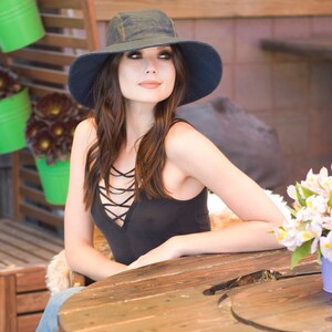 May include: A woman wearing a black tank top with a criss-cross design and a dark blue denim hat sits at a wooden table.