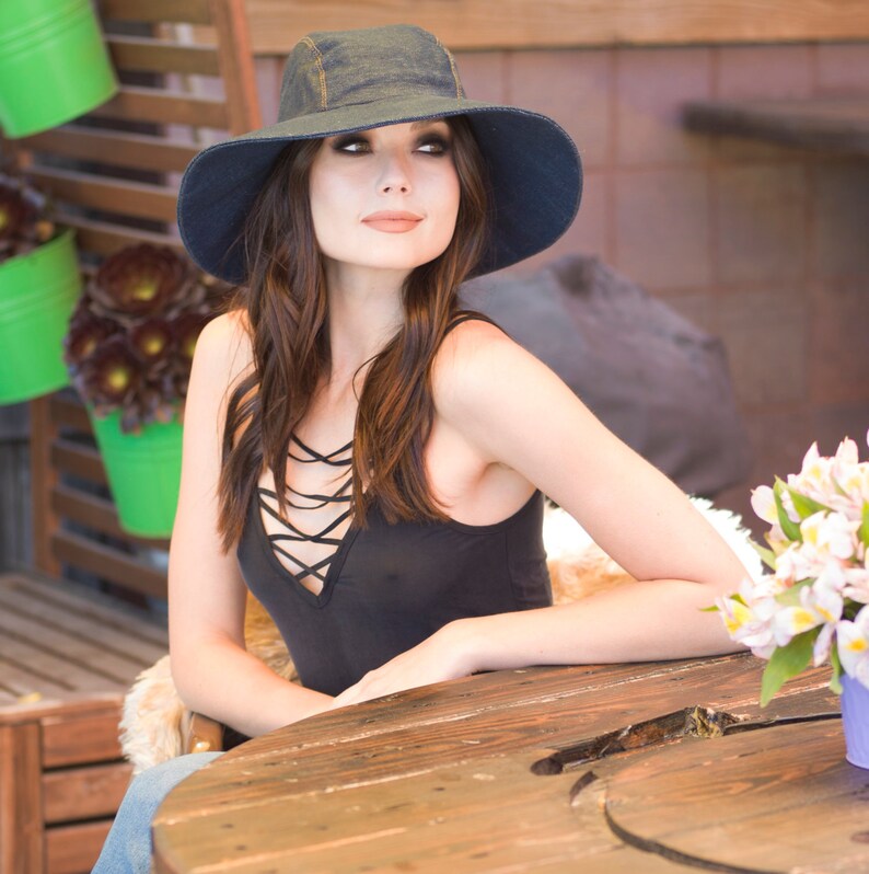 May include: A woman wearing a black tank top with a criss-cross design and a dark blue denim hat sits at a wooden table.